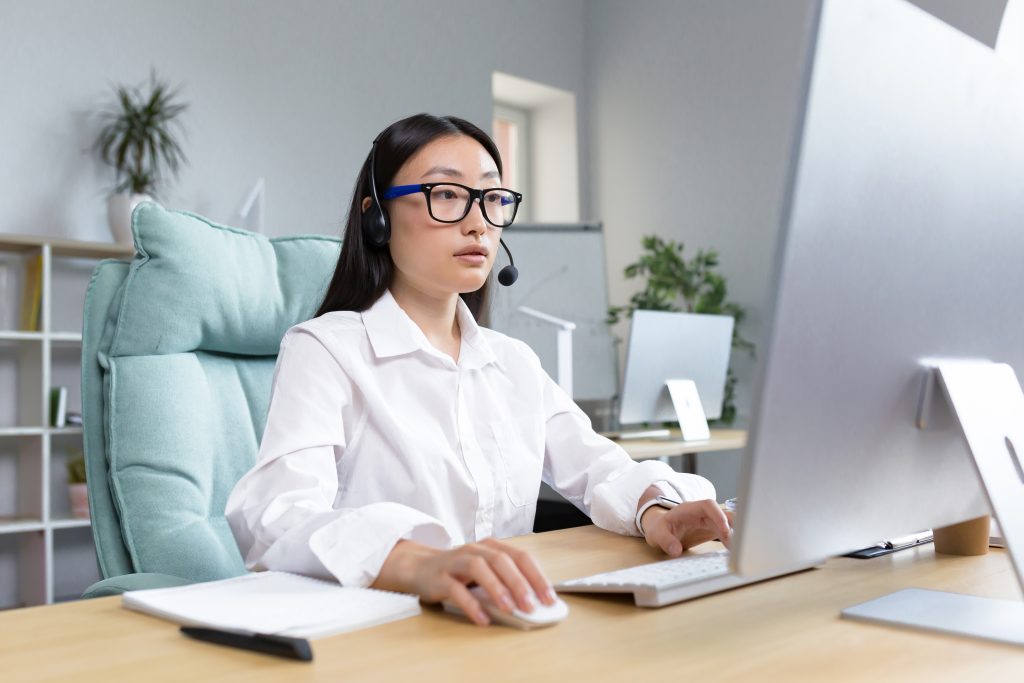 Online distance learning. Young beautiful Asian female teacher in headphones and glasses sitting at a table at a computer teaching a class lesson online.