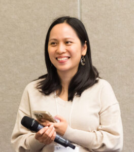 a professional headshot of Gwen Nguyen, a woman with long black hair, wearing a beige sweater, smiling while holding a microphone and a smartphone at a conference