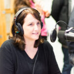 A photo of a woman with headphones on in front of a podcasting microphone.
