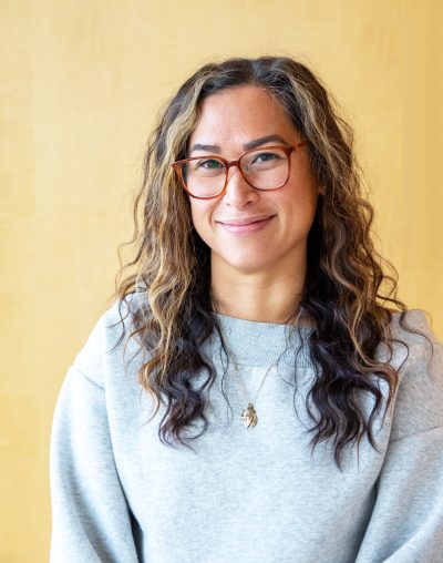 Headshot of Albertine De Leon in a grey sweatshirt on a yellow background, with glasses on!