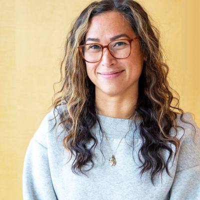 Headshot of Albertine De Leon in a grey sweatshirt on a yellow background, with glasses on!